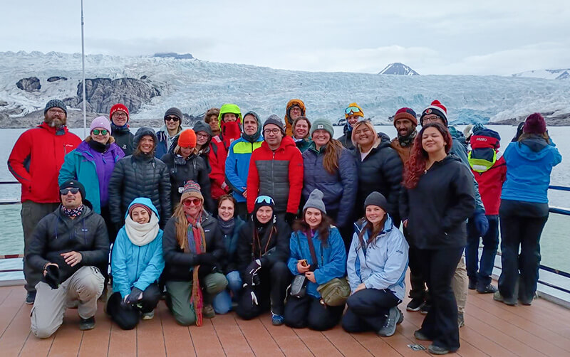 A group of college students and their professors, wearing cold-weather clothes, pose for a photo in front of a glacier in Svalbard.