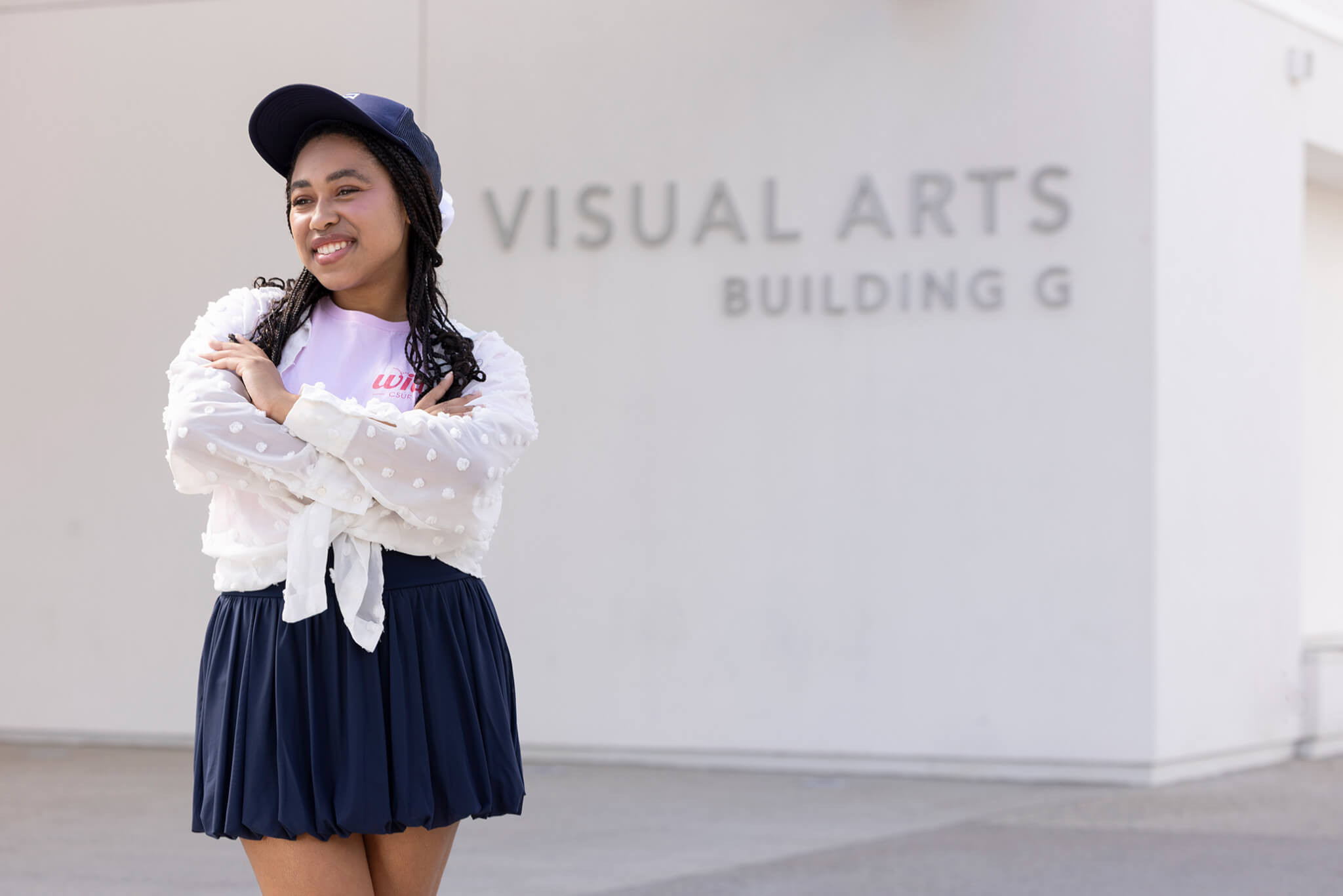 Skylar Williams standing outside the Visual Arts Building G, smiling with arms crossed, wearing a navy skirt, white textured blouse, and navy cap.