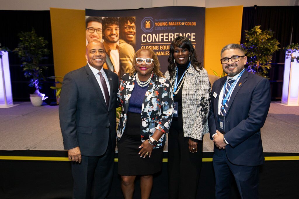 Four people stand together smiling in front of a banner that reads “California State University Young Males of Color Consortium Conference.”