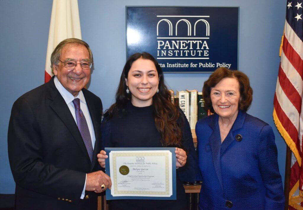 Leon Panetta, Melyne Garcia and Sylvia Panetta hold a certificate for the 2025 Panetta Congressional Internship, while smiling for a group photo in front of a sign for the Panetta Institute for Public Policy.