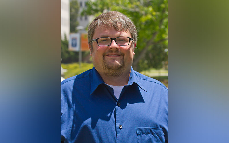 Man wearing glasses and a blue button-down shirt, smiling outdoors with trees and campus buildings in the background.