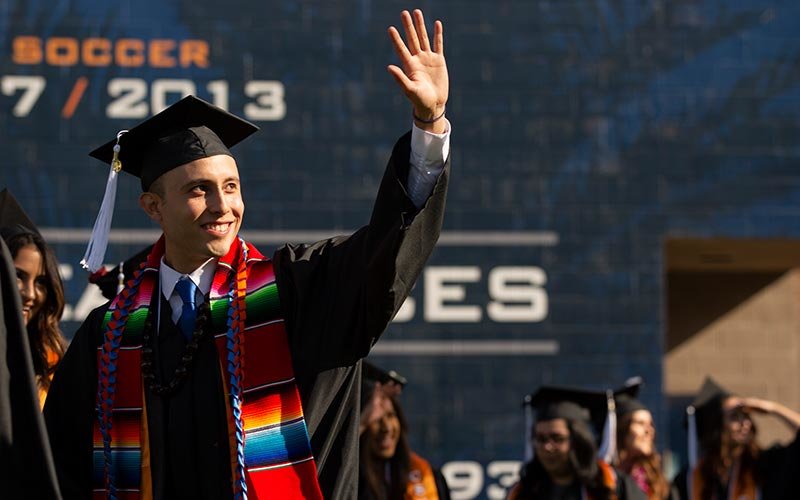 Hispanic graduate waves to his family in the stadium.