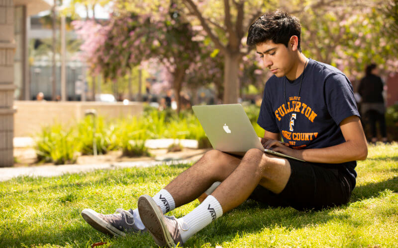 Student seated on grass working on laptop computer.