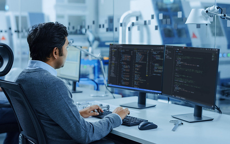 Man wearing a blue sweater and blue collared shirt sitting at a desk doing software, application, security development/engineering work on two monitors