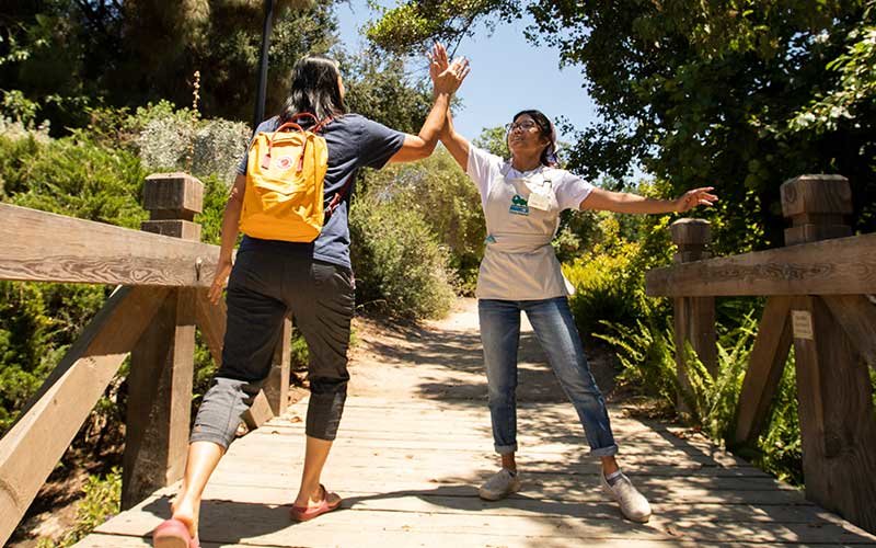 Visitor wearing navy blue t-shirt, black capris, sandals, and a backpack high-fives a Fullerton Arboretum greeter wearing a white t-shirt, blue jeans, sneakers and an apron, crossing the bridge into the gardens.