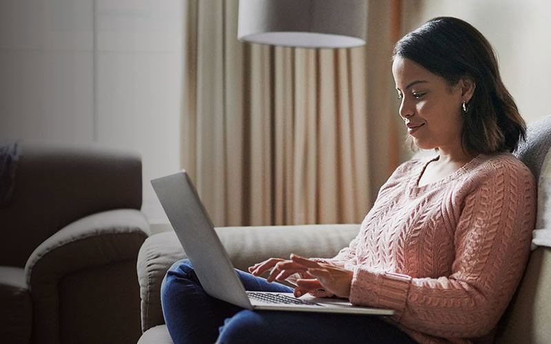 Woman sitting on a gray or tan fabric sofa wearing denim jeans and a pink sweater with a laptop on her lap.