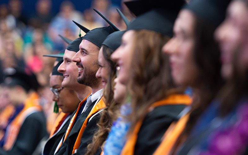 Diverse group of 8 CSUF graduates sitting at the graduation ceremony in their cap and gown, smiling.