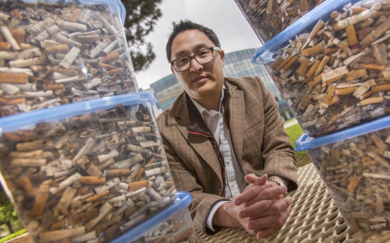 Asian man in sport coat sits between two stacks of plastic containers holding cigarette butts.