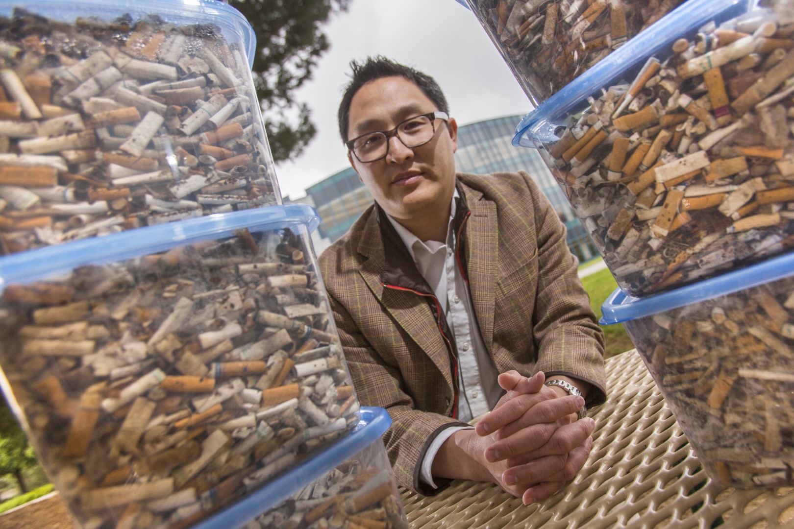 Asian man in sport coat sits between two stacks of plastic containers holding cigarette butts.