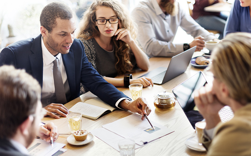 Male in dark suit, gray tie presenting ideas to three people while a female coworker is supporting him with a laptop..