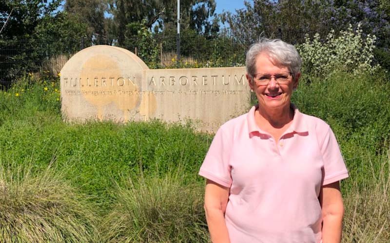 Female wearing pink polo in front of the Fullerton Arboretum sign that is surrounded by various green plants and trees