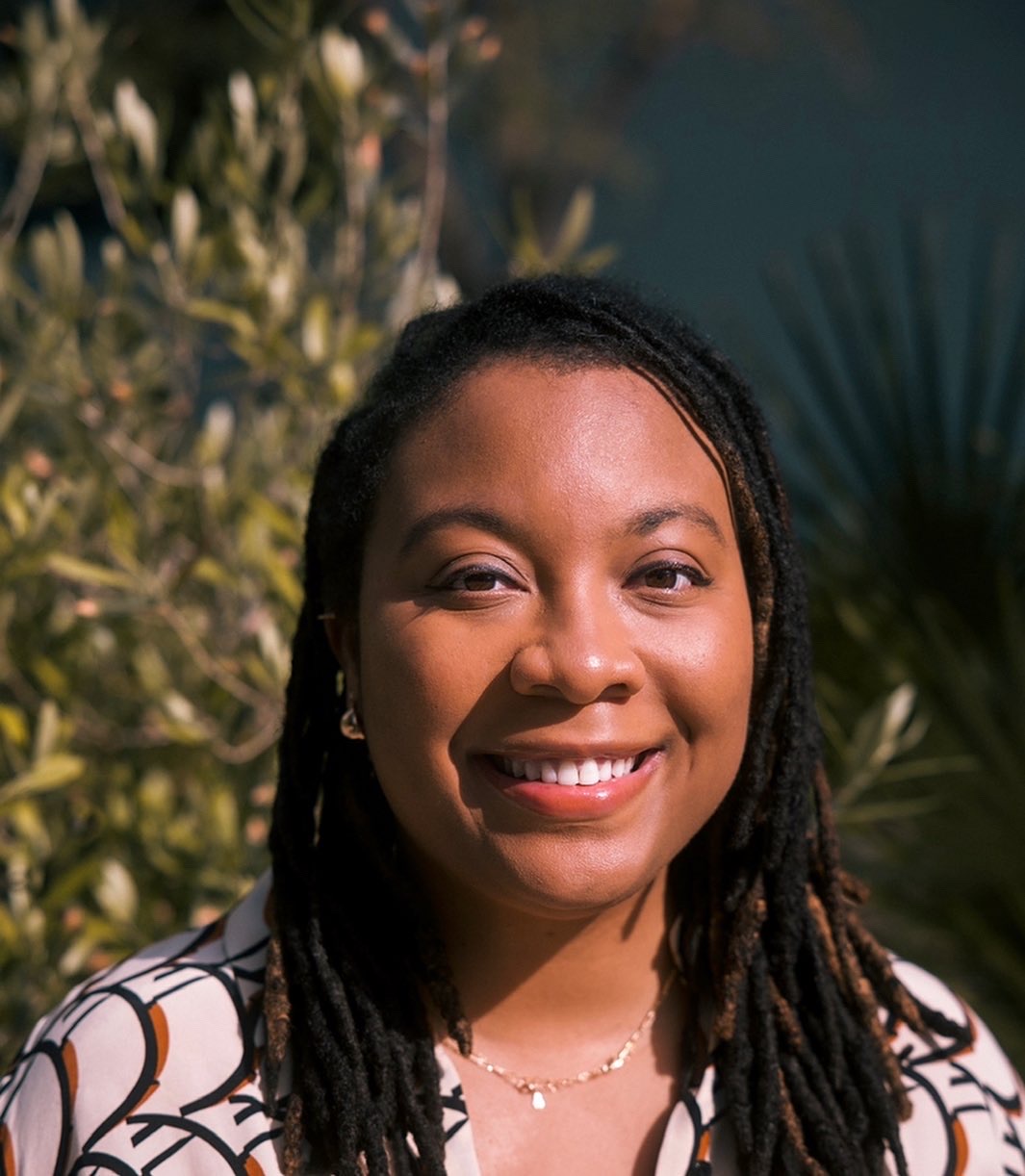 Headshot of Kristin Rowe. She has shoulder length locs and wears a tan printed shirt. She smiles, and greenery is blurred in the background.