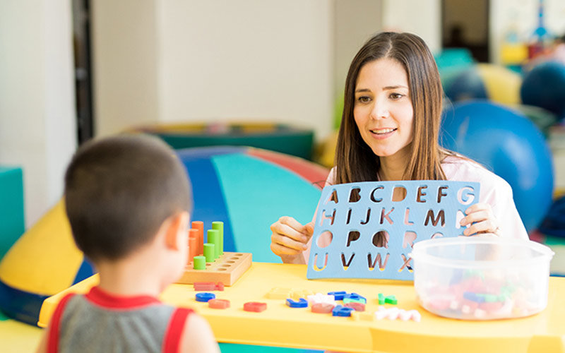 Teacher holds a puzzle, missing the letters of the alphabet, in front of a child, with the letter puzzle pieces on the table.