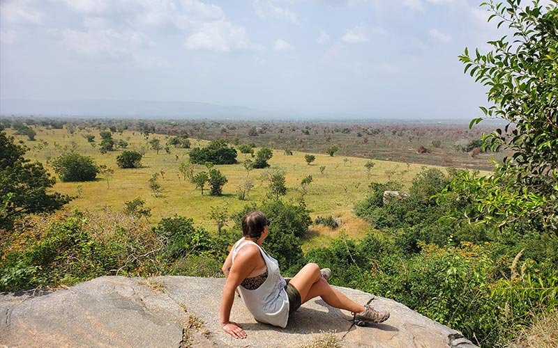 Student wearing a tank top, shorts and hiking shoes, sitting on a rock formation looking out into the African landscape filled with trees and grass.