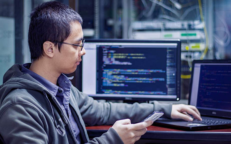 Young man with glasses, collared shirt and jacket working on his mobile phone, laptop, and desktop to secure his organization's network