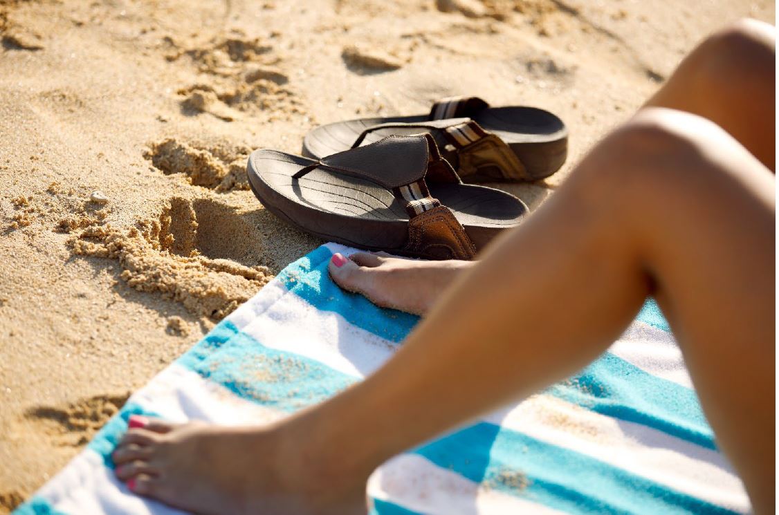 Woman laying on a beach towel on the sand, with sandals next to her