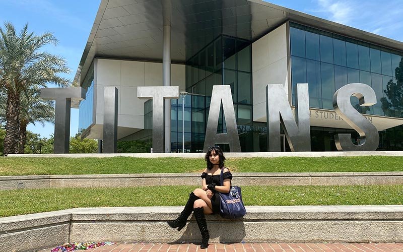 Female student in a black dress seated on the steps in front of oversized letters that spell out TITANS in capital letters on the campus of Cal State Fullerton (CSUF)