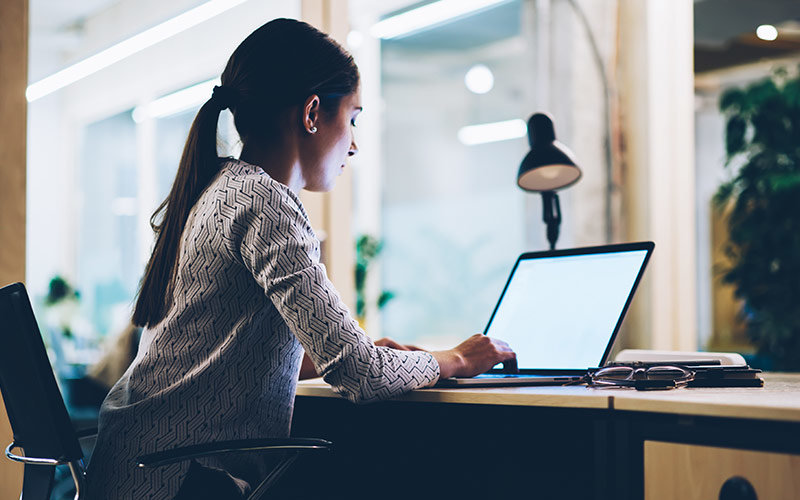 Female with a long ponytail and a long-sleeve shirt working on her laptop at a desk.