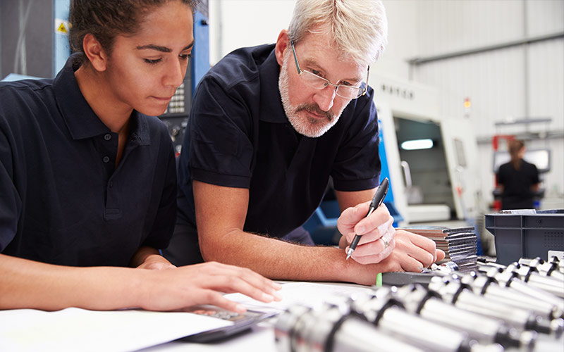 Female worker, hair pulled back, in a navy polo working alongside an older male worker in a navy polo as they review work
