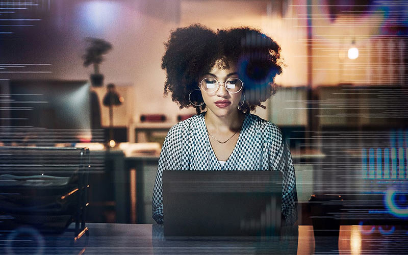 African American woman, wearing glasses, hoop earrings, necklace and geometric patterned top, sitting at desk and working on laptop