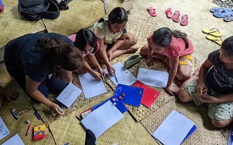 Dark-haired female adult kneeling down to help four children, seated or kneeling, with learning exercises.