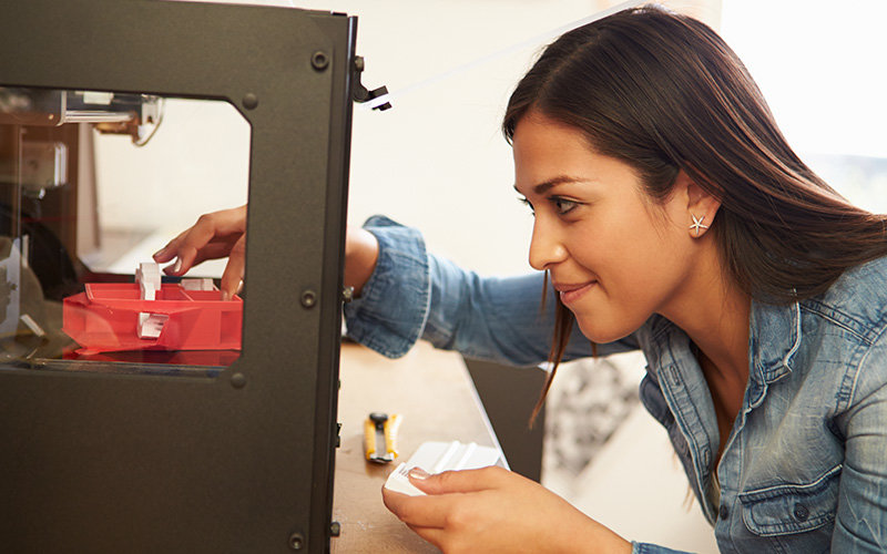 female with long dark hair, silver star earrings, and wearing a blue long-sleeve denim shirt working on a 3D printer