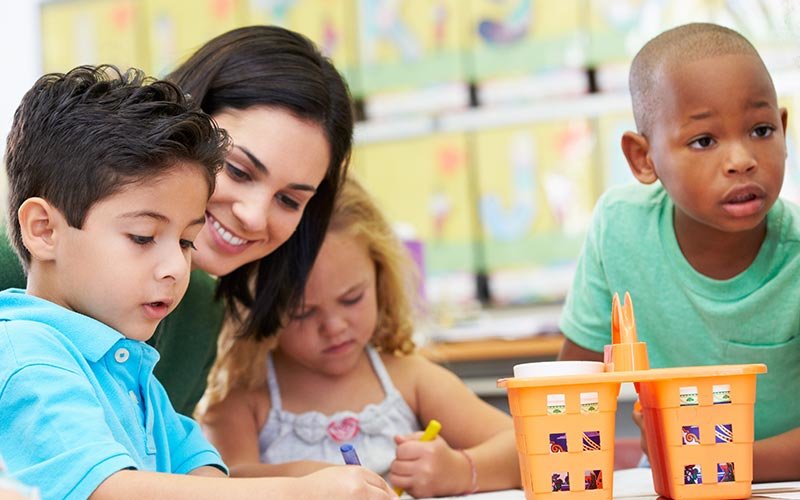 Female teacher with dark hair happily working with three students - dark-haired boy in a teal polo shirt drawing with a blue crayon, girl in a white top coloring with a yellow crayon and an African-American boy in a green shirt