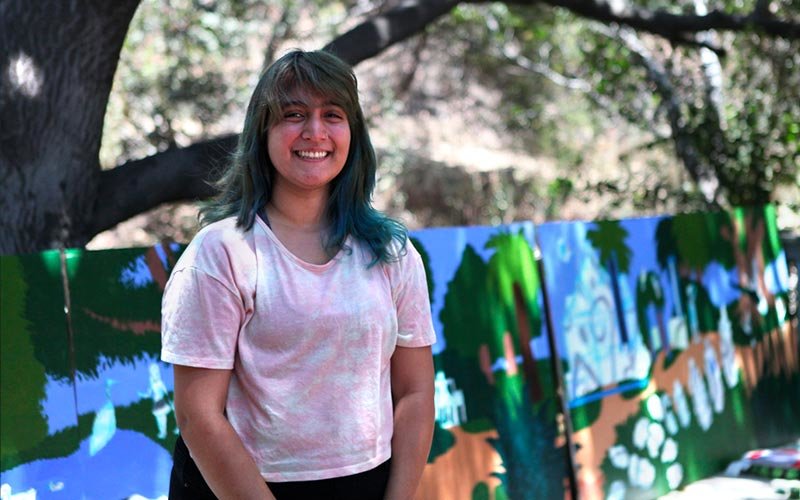 Female with a pink shirt standing in front of a work-in-progress mural of the Fullerton Arboretum