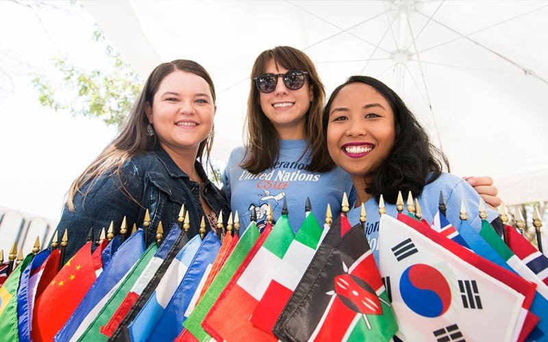 Three diverse females all with dark hair standing behind miniature flags of the world.