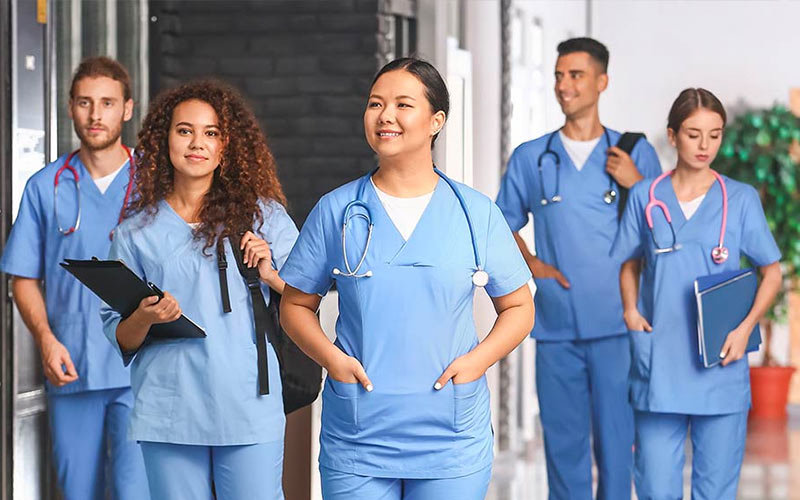 Five diverse medical students in blue scrubs - male with short brown hair and facial hair with a stethoscope, female with curly brown hair with a backpack and clipboard, female with with pulled back black hair and stethoscope, female with pulled back brown hair with files and stethoscope, male with short dark hair with backpack and stethoscope
