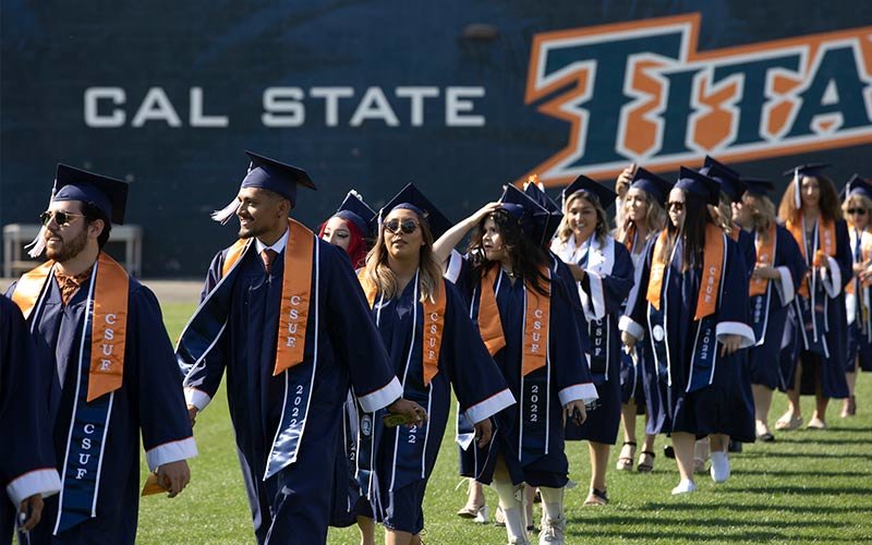 Single-file line of Cal State Fullerton graduates, both males and females, in their navy blue graduation cap and gown in front of signage that reads Cal State Fullerton Titans