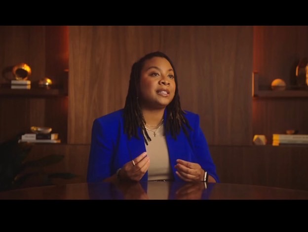 Kristin Rowe being interviewed in documentary. She sits at a wooden table, and she wears a blue blazer. She has locs and make up on. She is in the middle of speaking, with her hands gesturing.