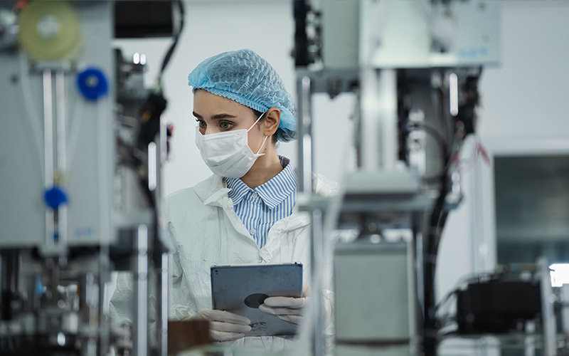 Person in a medical facility observing equipment and taking notes on a tablet while wearing a hairnet, white jacket and gloves.