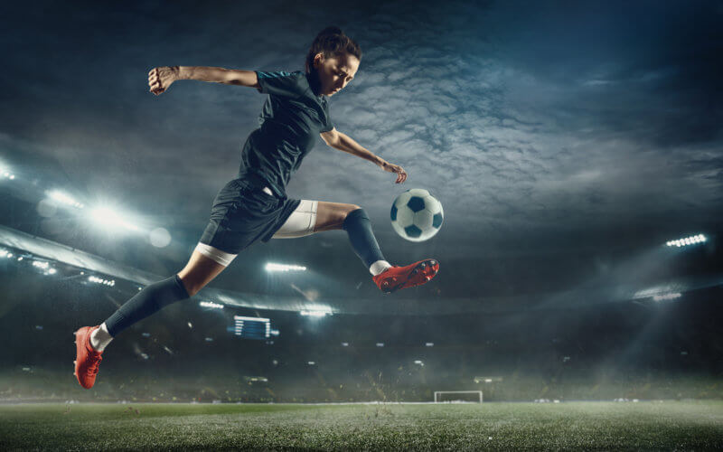 Young female soccer or football player with long hair in sportwear and boots kicking ball for the goal in jump at the stadium. Concept of healthy lifestyle, professional sport, hobby, motion, movement.