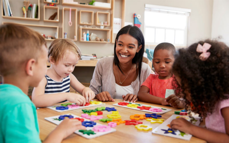 Preschool teacher sitting with four diverse children at a table as they work on a educational activity.
