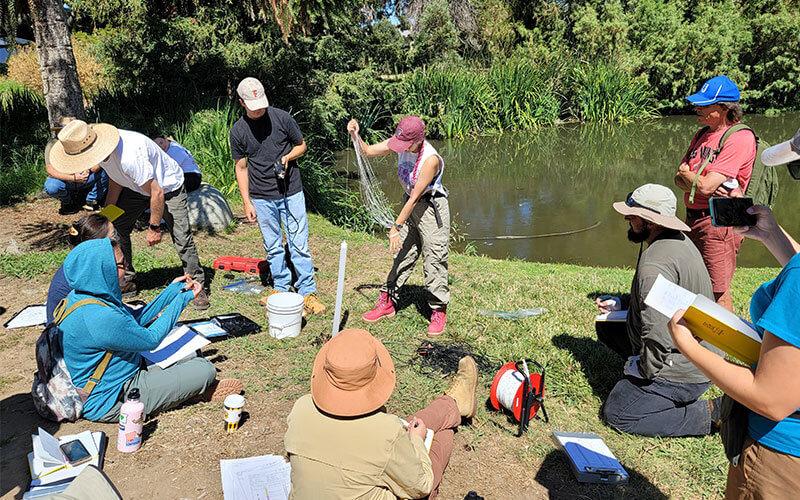 Students gathered around the lake at Fullerton Arboretum working on a hydrology project. They have various tools including buckets, rope, and data charts.
