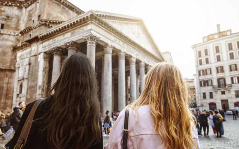 Two girls standing in front of the Pantheon in Rome. The girl on the left has dark brown hair and a black jacket. The girl on the right has blonde hair and a pink jacket. There are several other people gathered around the Pantheon.
