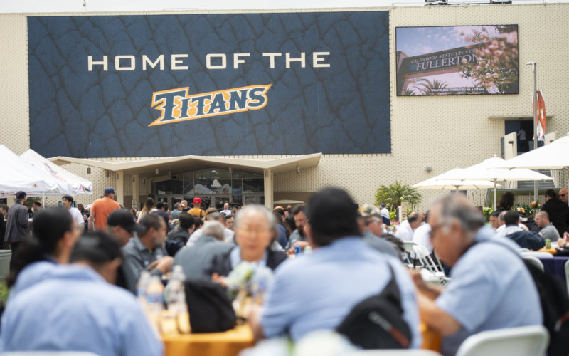 CSUF staff and faculty sit at round tables and some with umbrellas on a field under a large banner that says Home of the Titans.