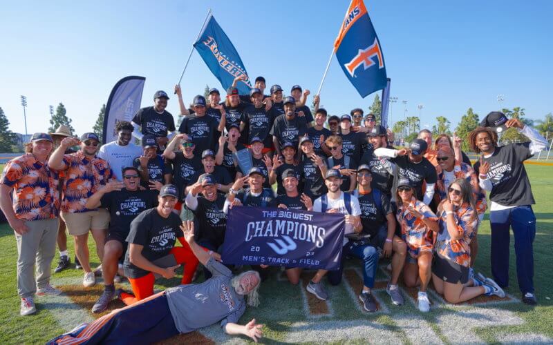 a group of men smiling waving a Big West Track championship banner