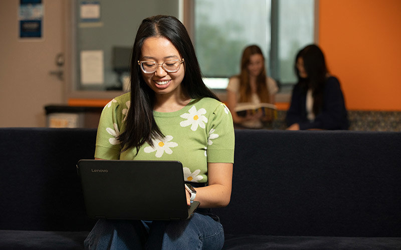 Young female student wearing glasses and a green floral shirt sitting in the CSUF library typing on a laptop.