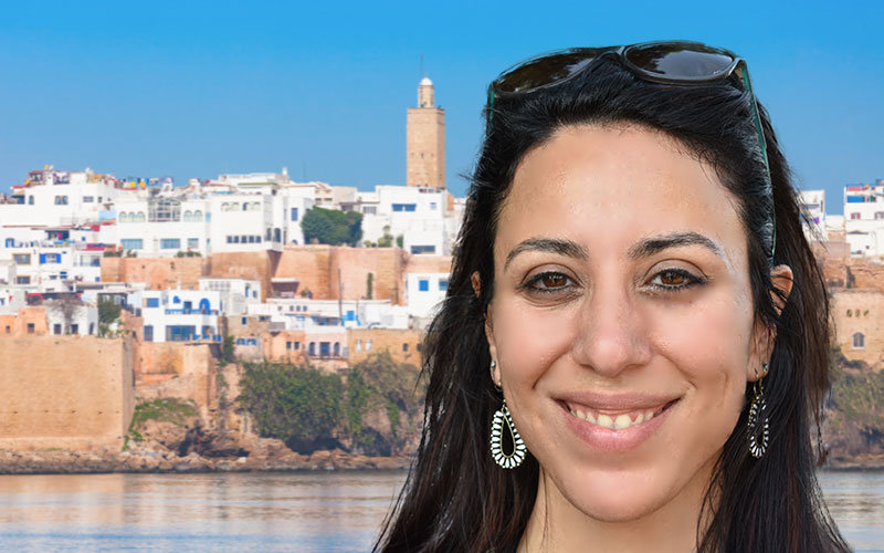 Headshot of Yasmine Shereen, photoshopped over a background of Rabat, Morocco. The picture of Rabat is taken on a sunny, cloudless day on the edge of the water. There are many white and brown buildings clustered on a small hill.
