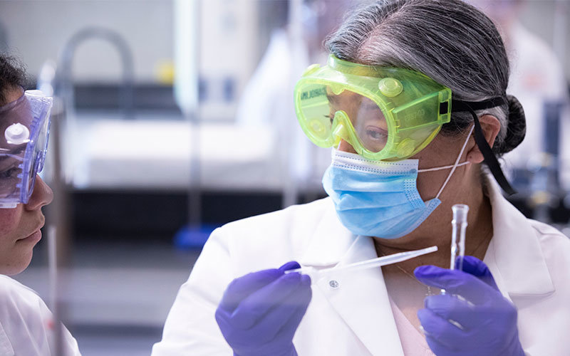 Two diverse females wearing lab coats and safety goggles. The female on the right is holding a dropper and test tube.