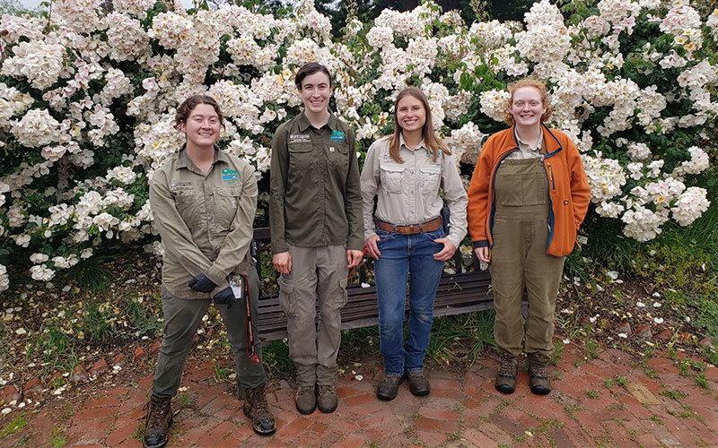 The four female Fullerton Arboretum horticulturists standing in front of a huge bush of white flowers. Pictured from left to right is Marjorie Rhodes, Amy Bulone, Anna Baier, and Mars Jordan.