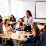 Female High School Teacher Standing By Student Table Teaching Lesson