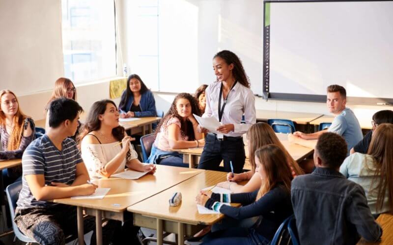 Female High School Teacher Standing By Student Table Teaching Lesson