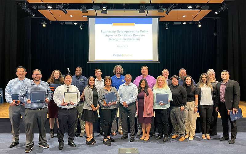 Twenty diverse male and female adults standing in a line, each holding up certificate. Behind them is a large projector screen which reads: "Leadership Development for public Agencies Certificate Program Recognition Ceremony, May 8, 2023.