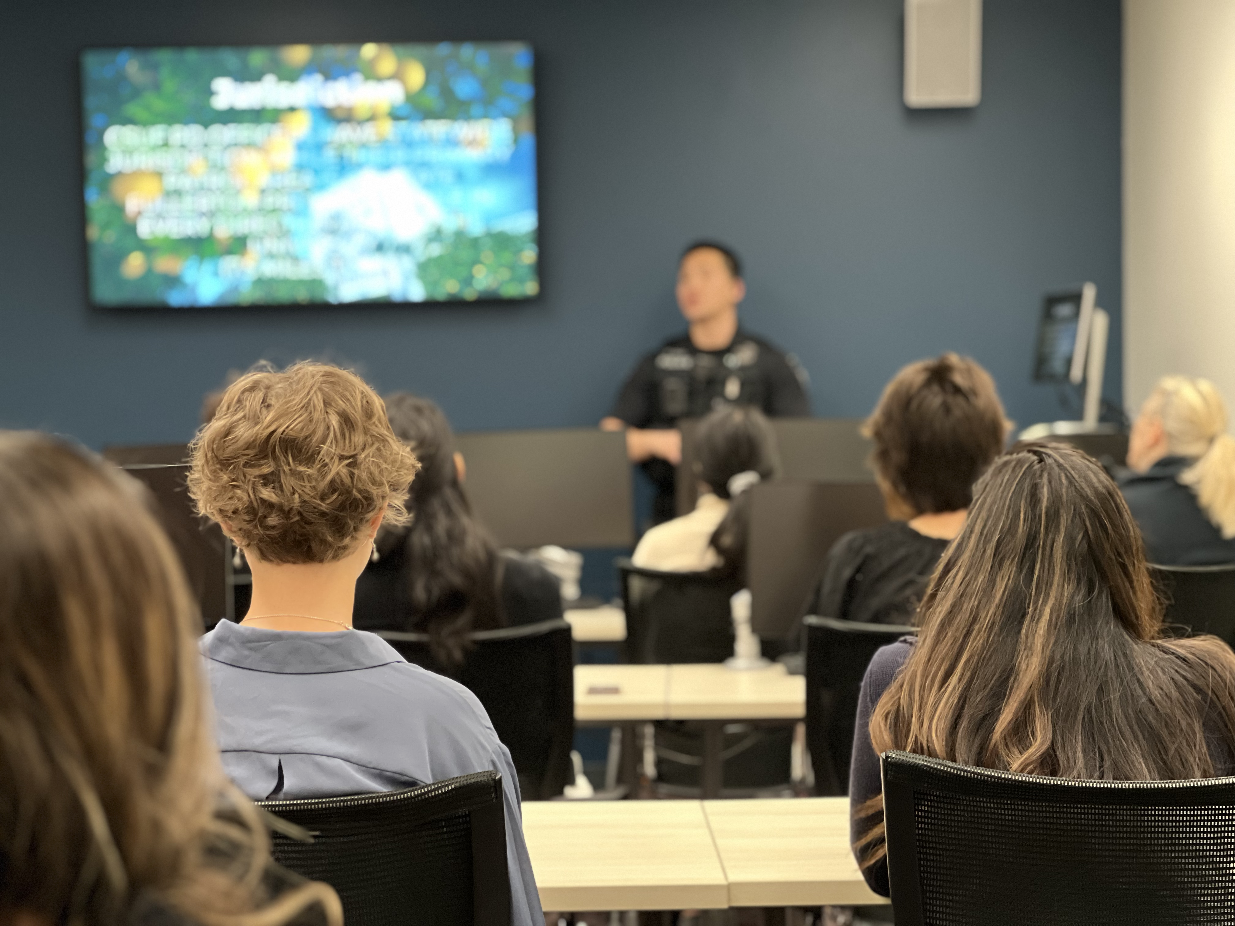 Multiple people sitting at a table with their backs to the camera. They are listening to a presentation from a police officer that is standing at the front of a room slightly blurred out. Slightly visible in the background is a TV monitor with the presentation they're hearing.