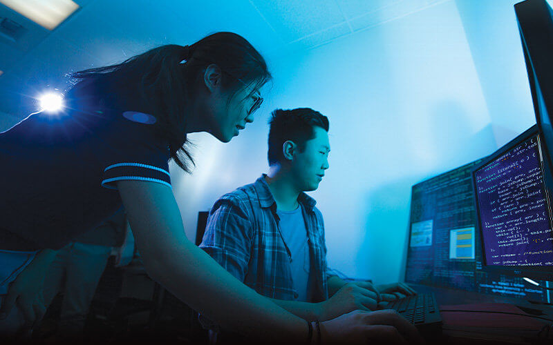Male and female student in a dimly lit classroom with blueish-green lighting. They are looking at three computer screens which display code.