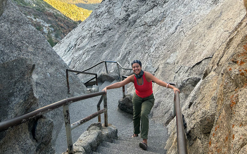 Delaney Cabral wearing hiking attire and climbing up a staircase made of stone that's built into a mountain. She's smiling at the camera and holding onto the metal railings with both hands. Behind her in the background is a view of lush green mountains, illuminated by the sunlight.