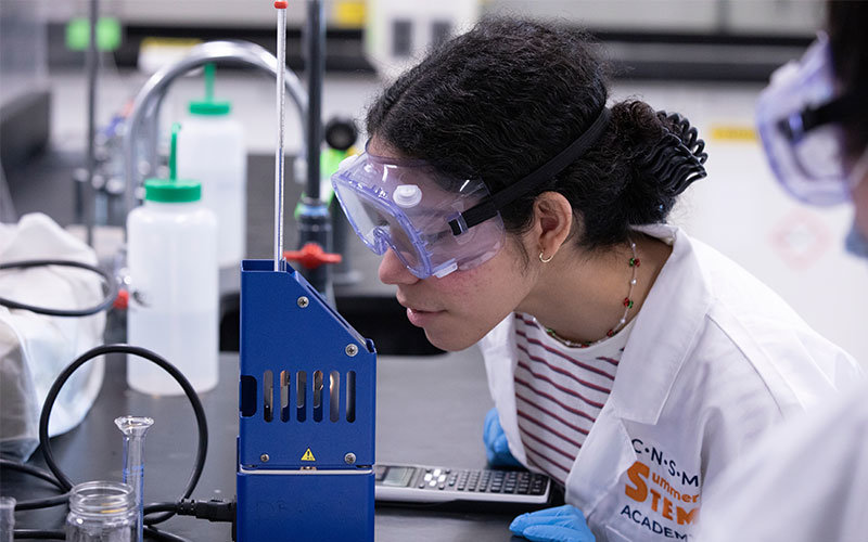 Diverse female wearing safety goggles and lab coat inspecting test tubes.